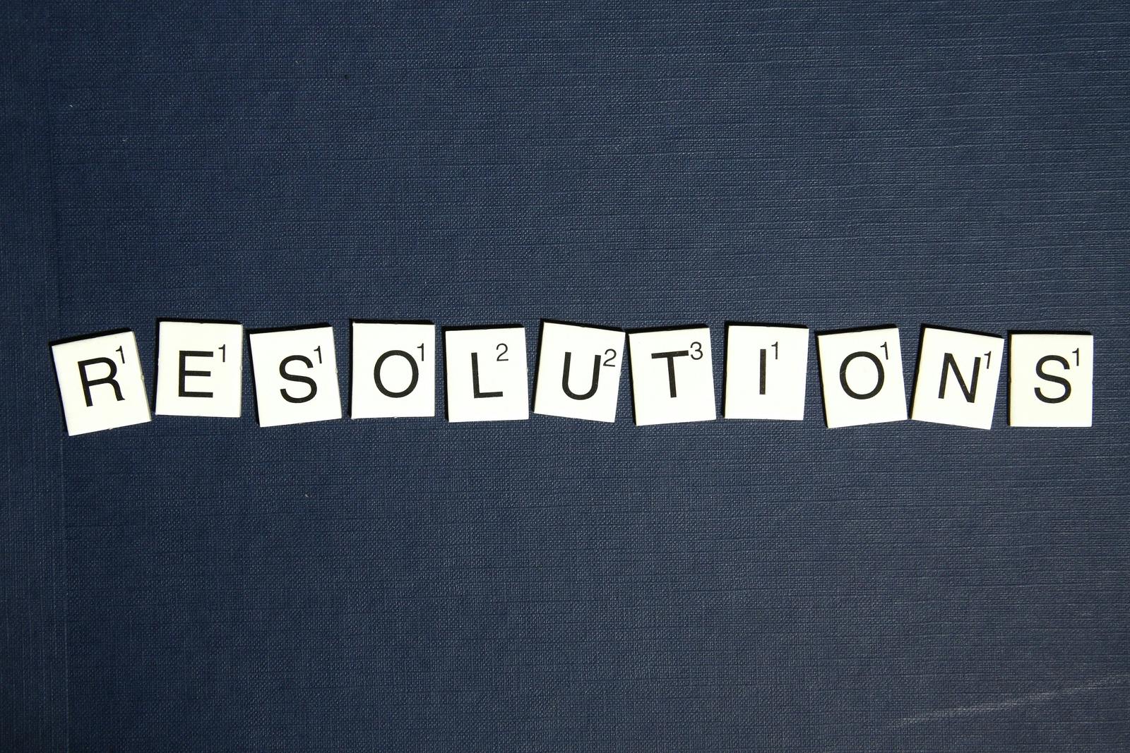 Scrabble tiles spell 'RESOLUTIONS' on a textured dark blue background, capturing a New Year theme.