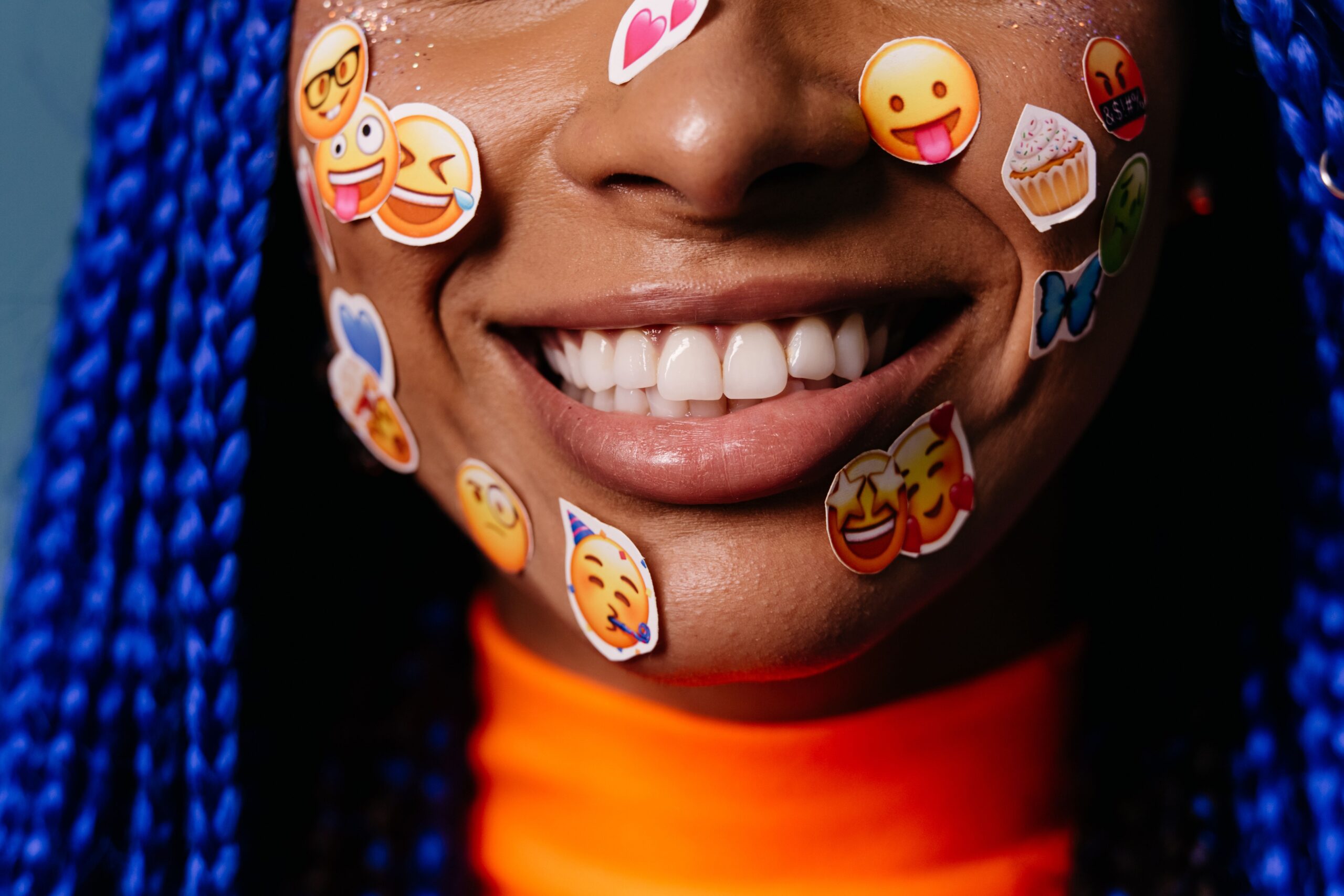 Close-up of a smiling woman with blue hair and emoji stickers on her face, expressing joy.
